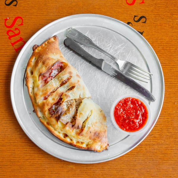 A calzone with meat and cheese, served with a side of marinara sauce on a metal tray.