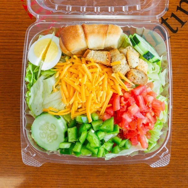 A fresh garden salad in a clear plastic container with lettuce, cheese, tomatoes, cucumbers, and croutons.