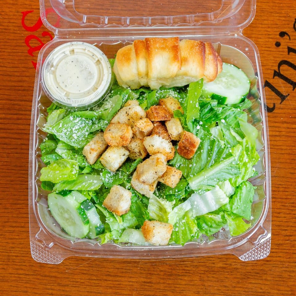 Caesar salad with croutons, cucumber, and a breadstick in a clear plastic container.