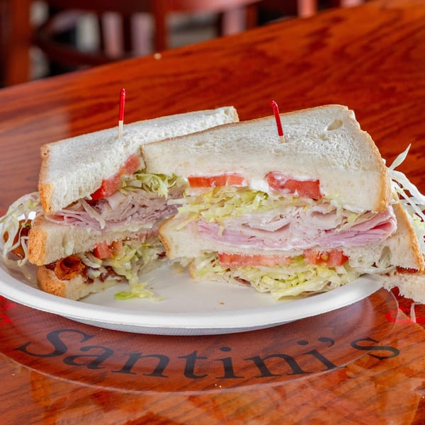 Stacked deli sandwiches with turkey, ham, lettuce, tomato, and mayonnaise on white bread, served on a plate.