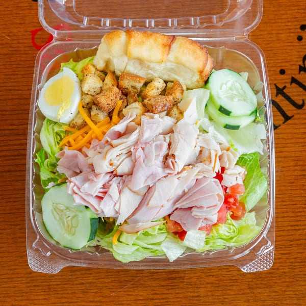 A clear plastic container holds a chef salad with ham, turkey, egg, croutons, and vegetables.