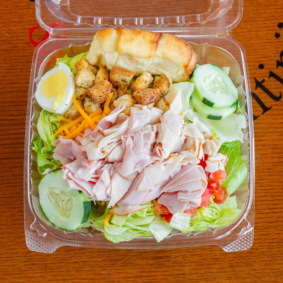 A clear plastic container holds a chef salad with ham, turkey, egg, croutons, and vegetables.
